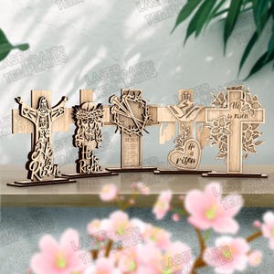 May include: A collection of five wooden crosses with intricate laser-cut designs. Each cross features religious imagery, including Jesus, a crown of thorns, and floral elements. The words "He is Risen" are displayed on some crosses. The crosses are displayed on a wooden surface.