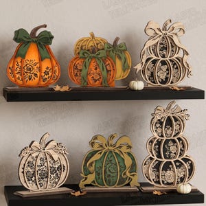 May include: A collection of six decorative wooden pumpkin cutouts displayed on two black shelves. The pumpkins feature intricate floral patterns, layered designs, and attached bows in shades of orange, green, black, and natural wood tones.