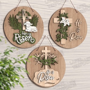 May include: Three wooden Easter decorations with the words "He is Risen". Each round sign features a cross, with additional designs including a lamb, a lily, and a crown of thorns. The signs are light brown with green and white accents.