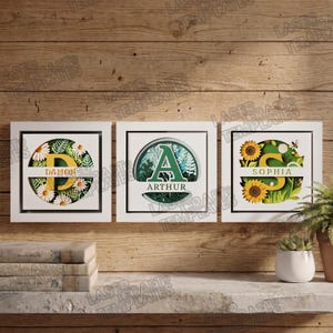 May include: Three framed wall art pieces with personalised initials and names. The first features a yellow "D" with daisies, the second a green "A" with foliage, and the third a yellow "S" with sunflowers. Each piece has a white frame.