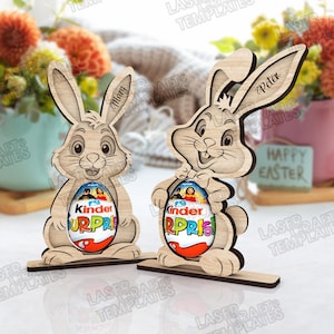 May include: Two wooden Easter bunny decorations, each holding a Kinder Surprise egg. One bunny is labeled "Mary" and the other "Peter". The eggs are red, white, and blue. The words "Happy Easter" are visible.