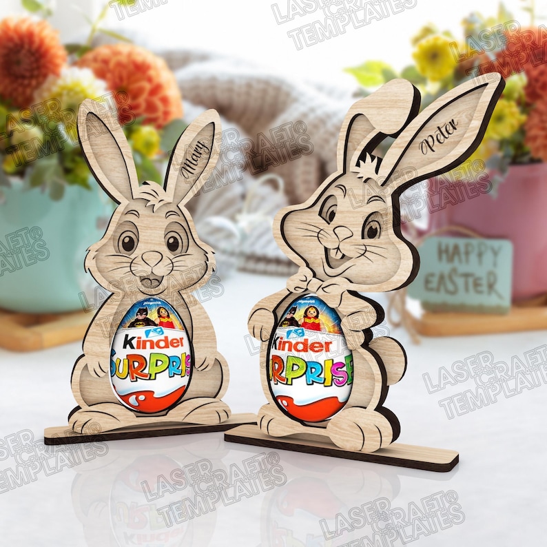 May include: Two wooden Easter bunny decorations, each holding a Kinder Surprise egg. One bunny is labeled "Mary" and the other "Peter". The eggs are red, white, and blue. The words "Happy Easter" are visible.