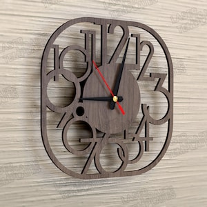 May include: Wooden wall clock with a dark brown finish. The clock has a unique design with cut-out numbers and a circular face. The clock hands are black, with a red second hand. The clock is mounted on a light wood background.