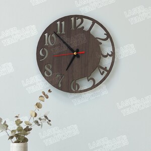 Puede incluir: Un reloj de pared redondo de madera con acabado marr&oacute;n oscuro. El reloj presenta n&uacute;meros recortados y un dise&ntilde;o decorativo en el lado derecho. Las manecillas son negras, con un segundero rojo. El reloj est&aacute; montado en una pared blanca. Di&aacute;metro: aprox. 30 cm.