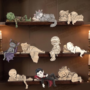 May include: A collection of nine wooden laser-cut figures depicting sleeping characters including a green zombie, a grey cat, a witch, a bat, an owl, a mummy, a ghost, a skeleton, a vampire, and a pumpkin, all arranged on three shelves.