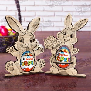 May include: Two wooden Easter bunny decorations. Each bunny holds a Kinder Surprise egg. One bunny has the name "Anna" on its ear, the other has "John". The bunnies are light brown and have a playful design.