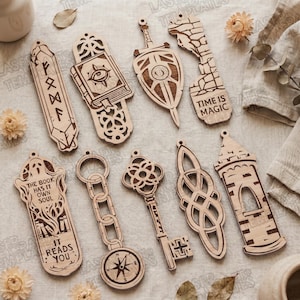 Op de afbeelding: Een verzameling van negen lasergesneden houten bladwijzers met ingewikkelde ontwerpen, waaronder een runenkristal, een spreukenboek, een zwaard, een stenen muur met de tekst TIME IS MAGIC, een geest met de tekst THE BOOK HAS IT OWN SOUL IT READS YOU, een ketting, een sleutel, een knoop en een toren.
