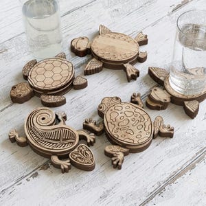 Puede incluir: Un juego de cuatro posavasos de madera con formas de animales, incluyendo una tortuga, una rana y un lagarto, con detalles grabados a láser. Dos vasos de cristal transparente se colocan sobre los posavasos de tortuga y rana, mostrando la veta de madera clara natural y los patrones grabados oscuros.