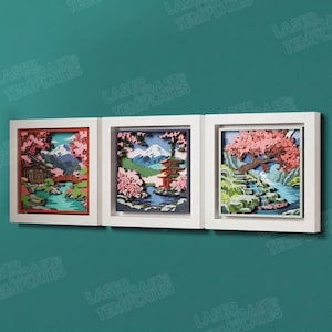 May include: A set of three square white-framed wall art pieces featuring layered laser-cut designs. Each frame displays a multi-dimensional landscape with pink cherry blossoms, blue water, green foliage, and structures like a pagoda and bridge.