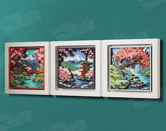 May include: A set of three square white-framed wall art pieces featuring layered laser-cut designs. Each frame displays a multi-dimensional landscape with pink cherry blossoms, blue water, green foliage, and structures like a pagoda and bridge.
