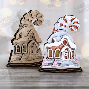May include: Two decorative wooden Christmas village houses. One is unfinished wood, the other is painted red, white, and pink with a snowy roof. Both have a whimsical, curved hat-like top and are on a wooden base.