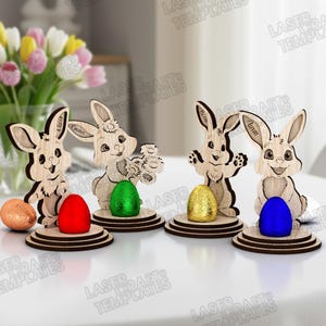 May include: Wooden Easter bunny decorations with colorful foil-wrapped eggs. Each bunny holds an egg or flowers, with names like "Oliver", "Emma", and "Aidan" engraved on them. The bunnies are on round wooden bases.