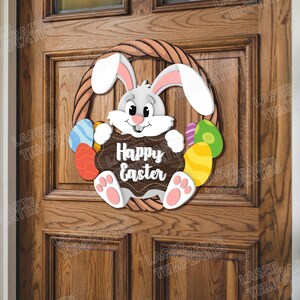 Pode incluir: Um coelho branco segurando um cartaz que diz "Feliz P&aacute;scoa" est&aacute; rodeado de ovos de P&aacute;scoa coloridos em uma porta de madeira marrom.
