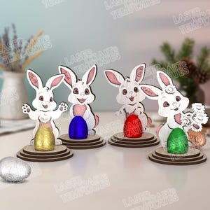 Peut inclure: Décorations de Pâques en forme de lapins en bois avec des œufs colorés. Chaque lapin blanc avec des accents roses tient un œuf de couleur différente. Les noms "Aleah", "Liam", "Chloe" et "Emma" sont écrits sur les lapins.