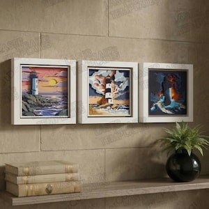 May include: Three framed lighthouse art pieces. Each piece features a layered paper design with a lighthouse, ocean, and sky. The frames are white, and the artwork is displayed on a textured wall. The lighthouses are in various colors.