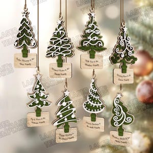 May include: A collection of wooden Christmas tree ornaments with intricate white designs and green bases. Each tree hangs from a string and has a small wooden sign with a holiday message and family name. Festive decor.