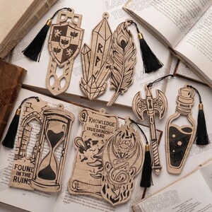 Op de afbeelding: Een verzameling van acht lasergesneden houten bladwijzers met ingewikkelde ontwerpen, waaronder een schild, kristallen, een veer, een zandloper, een perkamentrol met de tekst 'KNOWLEDGE IS THE TRUE DRAGON'S HOARD', een draak, een bijl en een flesje, elk met een zwart kwastje.