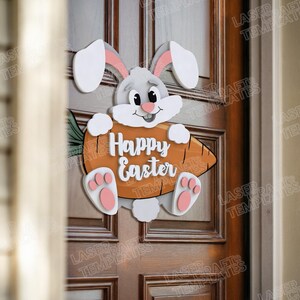 Pode incluir: Um coelho da P&aacute;scoa de madeira segurando uma cenoura com o texto "Happy Easter" nela. O coelho &eacute; branco com pelo cinza e orelhas e patas cor-de-rosa. A cenoura &eacute; laranja e tem folhas verdes.