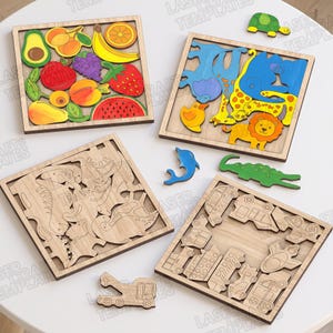Peut inclure: Quatre plateaux de puzzle carrés en bois avec des motifs découpés au laser. Un plateau contient des formes de fruits colorés, un autre des figurines d'animaux vibrantes, un troisième des contours de dinosaures et le quatrième des silhouettes de véhicules.