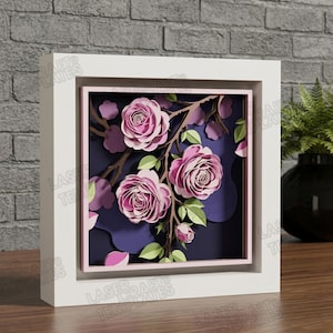 Puede incluir: Una obra de arte en papel enmarcada con rosas rosas en capas sobre una rama con hojas verdes, sobre un fondo azul oscuro. La obra de arte está encerrada en un marco blanco, creando un efecto 3D.