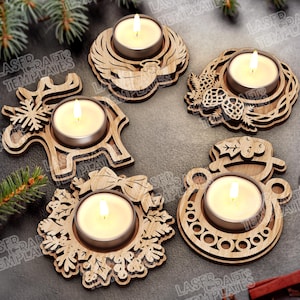 May include: A collection of five wooden tea light candle holders with festive designs. Each holder features a different holiday-themed shape, including a reindeer, angel wings, a wreath, a pinecone, and a Santa Claus ornament. Each holder contains a lit tea light candle.