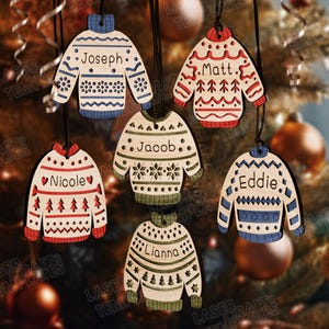 May include: Wooden sweater-shaped Christmas ornaments with names like "Joseph," "Matt," and "Nicole." Each ornament features a unique design with red, blue, and green accents. The ornaments are hanging in front of a blurred Christmas tree background.