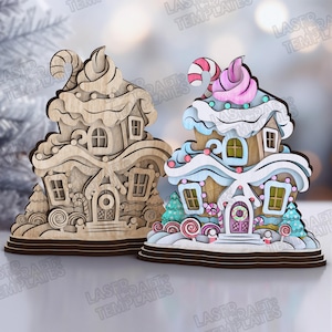 May include: Two gingerbread house ornaments. One is unpainted wood, the other is painted with pastel colors, candy accents, and a pink swirl on top. Both have windows, doors, and candy cane details. The ornaments are on a brown base.