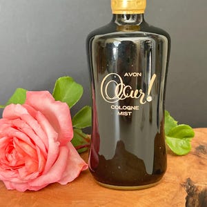 Op de afbeelding: Een zwarte glazen fles Avon Occur! Cologne Mist met een goudkleurige dop en een verstuiver. De voorzijde van de fles is voorzien van gouden letters en een kleine ingebedde strass-steen. Een enkele roze roos met groene bladeren staat links.