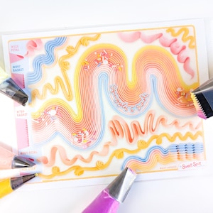 May include: A clear plastic stencil with colorful frosting designs in pink, yellow, and blue, with the text "Squiggles & Swirls." Several piping tips in various colors surround the stencil, including pink, yellow, and purple.
