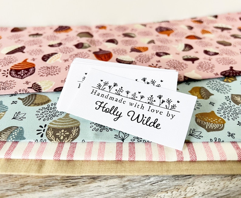 Puede incluir: Etiqueta blanca con texto negro que dice "Handmade with love by Holly Wilde" sobre una pila de tela con un patr&oacute;n rosa, azul y blanco.