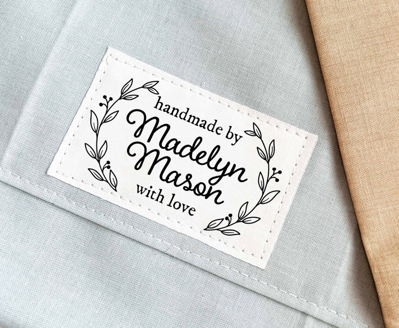 Puede incluir: Etiqueta de tela blanca con costuras negras alrededor de los bordes. La etiqueta tiene una corona floral negra con el texto "handmade by Madelyn Mason with love" en su interior.