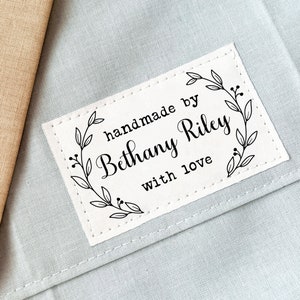 Puede incluir: Etiqueta de tela blanca con costuras negras alrededor de los bordes. La etiqueta tiene un dise&ntilde;o floral y el texto "handmade by Bethany Riley with love".