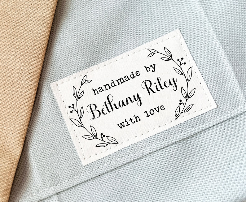 Puede incluir: Etiqueta de tela blanca con costuras negras alrededor de los bordes. La etiqueta tiene un dise&ntilde;o floral y el texto "handmade by Bethany Riley with love".