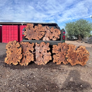 May include: Seven large, irregular wood slabs with prominent, swirling grain patterns and deep brown and amber tones. Two slabs are positioned in the bed of a dark green pickup truck, while five additional slabs are arranged upright on the ground.