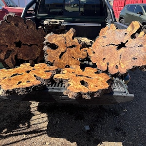 May include: Five large, irregular olive wood slabs with natural live edges and prominent grain patterns. Each slab features a dark, hollow center and a textured, light brown and golden yellow surface, displayed in the bed of a black pickup truck.