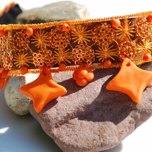 Puede incluir: Un collar gargantilla de tela marrón con abalorios en forma de estrella naranja y lentejuelas doradas.