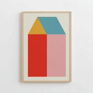 May include: Framed art print featuring a geometric house design. The house is divided into sections of red, pink, yellow, and blue. The artwork is framed in a light wood frame and set against a cream-colored background.