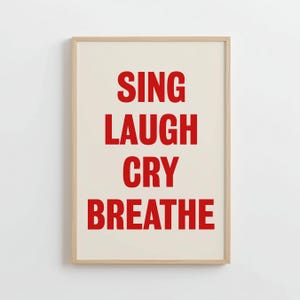 Puede incluir: Impresión enmarcada con las palabras "SING LAUGH CRY BREATHE" en letras rojas sobre un fondo crema. El marco es de color madera clara, con un diseño minimalista y moderno.