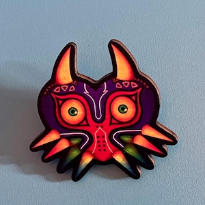 Könnte beinhalten: Eine dekorative Anstecknadel in Form einer stilisierten Maske mit violettem Gesicht, großen gelben Augen mit grünen Pupillen und orangefarbenen hornartigen Vorsprüngen. Das Design zeigt rote Akzente, weiße Linien und grüne Schattierungen.