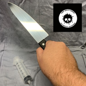 May include: A stainless steel chef's knife with a black handle is held in a hand. A syringe with a clear tube is connected to the knife. A black and white logo with a skull and the text "Aunt Dolly's Garage Los Angeles California" is in the background.