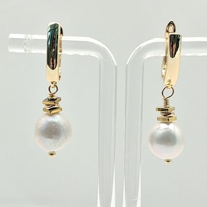 May include: A pair of gold-colored hoop earrings with a large, white, pearl-shaped drop. The earrings have a geometric design element and are displayed on a clear stand. The earrings are designed for pierced ears.