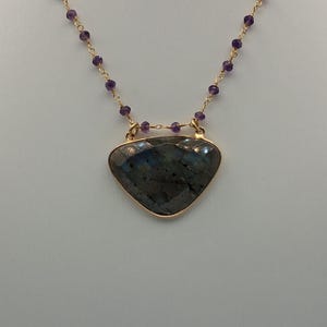 May include: A gold-toned necklace featuring a large, triangular labradorite pendant with a faceted surface. The pendant is framed in gold and is suspended from a chain of small, purple beads and gold links. The necklace is displayed against a white background.