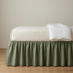 May include: A sage green bed skirt with a gathered ruffle design, positioned beneath a cream-colored mattress and a white folded blanket. The fabric features a visible texture, and the skirt extends to the floor, covering the base of the bed frame.