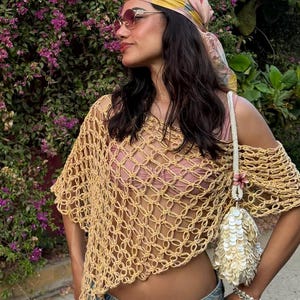 May include: A woman wearing a beige crochet top, blue jeans, and a floral scarf. She is also wearing sunglasses and carrying a white beaded purse with a flower accent. The background features purple flowers and green foliage.