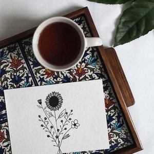 May include: A white ceramic teacup with a black floral design sits on a wooden tray with a colourful tile pattern. A white card with a black line drawing of a sunflower and other flowers is on the tray.