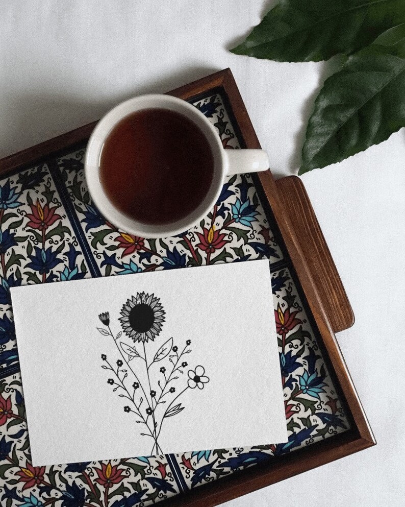 May include: A white ceramic teacup with a black floral design sits on a wooden tray with a colourful tile pattern. A white card with a black line drawing of a sunflower and other flowers is on the tray.
