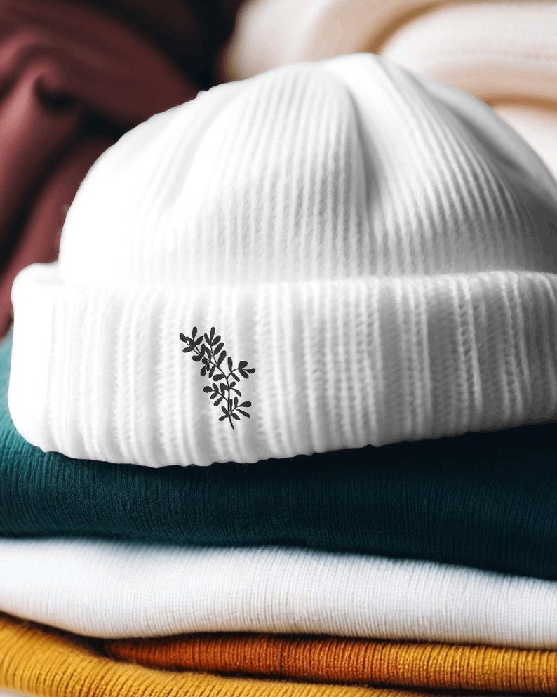 May include: White knitted beanie with a small black floral design on the front. The beanie is sitting on top of a stack of folded fabric in various colours.
