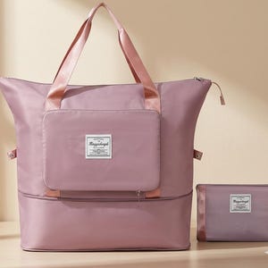 Puede incluir: Un bolso de viaje de color malva con asas a juego y un bolsillo frontal. Una bolsa plegable más pequeña está al lado del bolso. Ambas bolsas tienen una etiqueta con texto. El bolso está abierto y listo para usar.