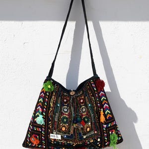 May include: Black shoulder bag with colorful embroidery, mirrors, and tassels. The bag features a geometric pattern and a wooden button closure. The bag is hanging against a white wall.