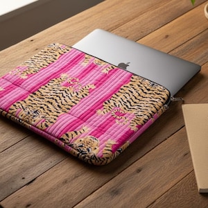 May include: A laptop computer is partially inserted into a pink and tan patterned laptop sleeve. The sleeve has a quilted design with vertical stripes and a tiger stripe pattern. A notebook and pen are on the wooden surface.